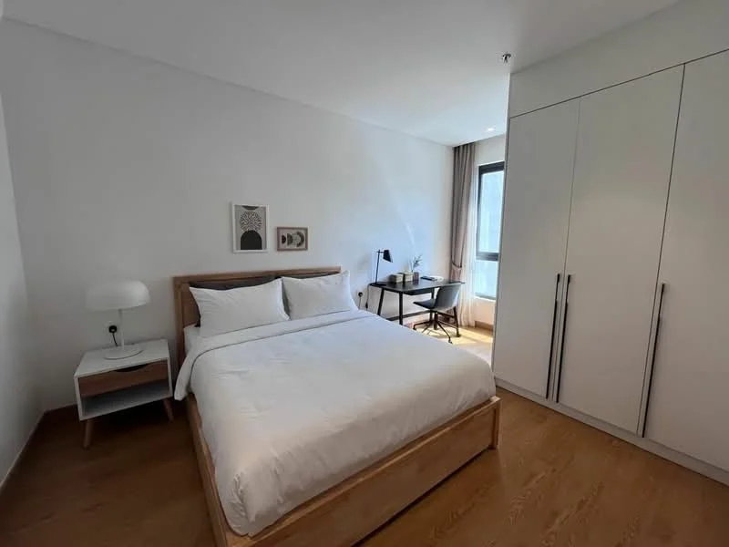 Bedroom features: queen-size bed with white bedding, wooden bed frame, nightstand with lamp, built-in wardrobe with white doors, study desk with chair, window with sheer curtains, wooden flooring, framed artwork on wall; style: modern.