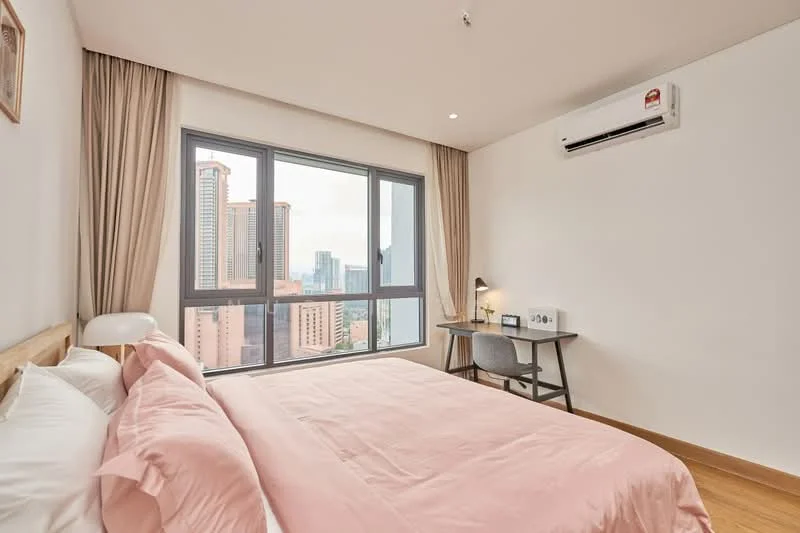 Bedroom features: king-size bed with pink bedding, wooden bed frame, floor-to-ceiling windows, city skyline view, study desk with lamp, grey chair, wall-mounted air conditioning unit, light wood flooring, curtains, decorative wall art; style: modern; view: city.