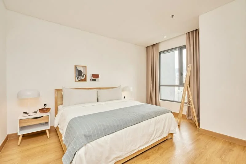 Bedroom features: king-size bed with white bedding, upholstered headboard, two bedside lamps, wooden nightstand, wooden flooring, floor-to-ceiling window, sheer curtains, decorative wall art, wooden ladder for decor, neutral painted walls; style: modern; view: city.