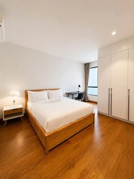 Bedroom features: king-size bed with wooden frame, white bedding, nightstand with lamp, built-in wardrobe with sliding doors, study desk with chair, window with natural light, wooden flooring, wall-mounted split AC, recessed ceiling lights, neutral painted walls; style: modern.