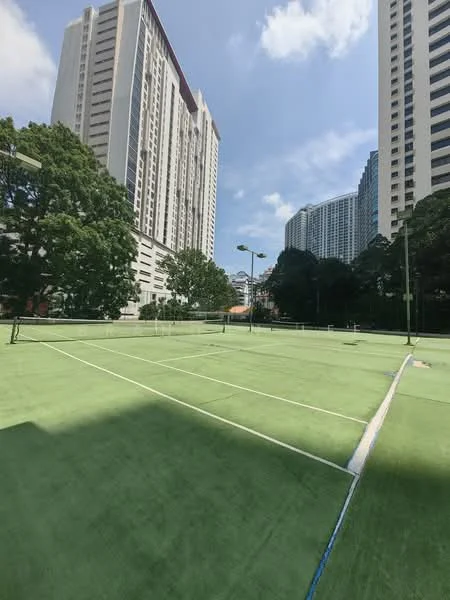Exterior type: tennis court; features: artificial grass surface, multiple tennis courts, surrounded by high-rise buildings, floodlights for night play, green landscaping, clear blue sky, urban setting; style: modern.