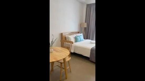 Bedroom features: queen-size bed with white bedding, two accent pillows in blue, wooden bed frame, side table with lamp, round wooden dining table, two wooden chairs, floor-to-ceiling window with grey curtains, light grey bedspread, neutral painted walls, tile flooring; style: modern.