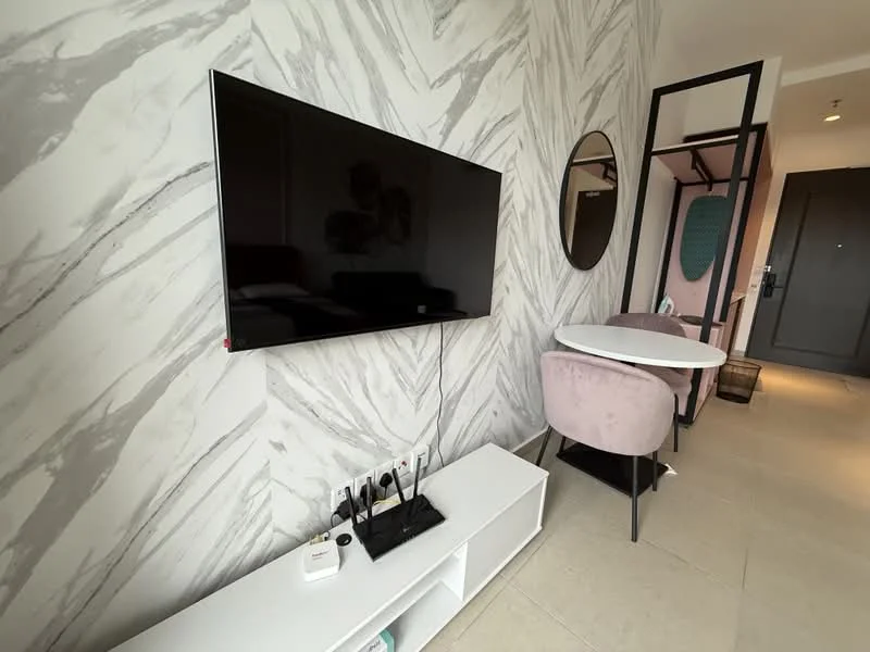 Living features: grey marble accent wall, white wall-mounted TV, white TV console, round mirror, oval dining table, pink upholstered dining chairs, floor tiles, door to hallway; style: modern.