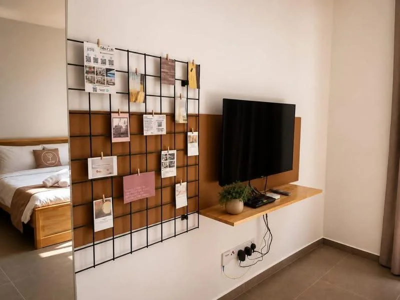 Room features: wall-mounted TV, wooden shelf for TV console, black metal grid wall with photos and notes, decorative plant on shelf, light-colored walls, grey tile flooring, curtains, bed visible in background with white bedding; style: modern.