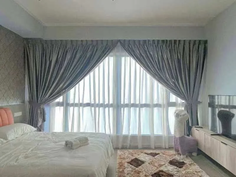 Bedroom features: queen-size bed with white bedding, grey upholstered headboard, floor-to-ceiling windows with sheer curtains, grey drapes, wooden flooring, decorative area rug with geometric pattern, wall-mounted TV, light wood bedside table, towel rolled on the bed, decorative plush llama, wall with textured wallpaper; style: modern.
