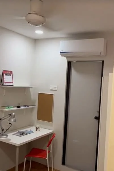Study features: white painted walls, wooden flooring, ceiling fan, wall-mounted split AC, desk with white surface, red chair, shelf with decorative items, framed wall calendar, corkboard, light fixture, door to hallway; style: modern.