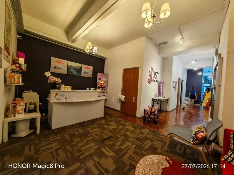 Room features: reception desk with white surface, wooden flooring, grey accent wall, decorative wall art, suspended ceiling with lights, two-tone wall paint, seating area with fabric chair, open layout, doorway leading to another room, countertop with coffee machine, shelves with decorative items, wall-mounted signage; style: modern.
