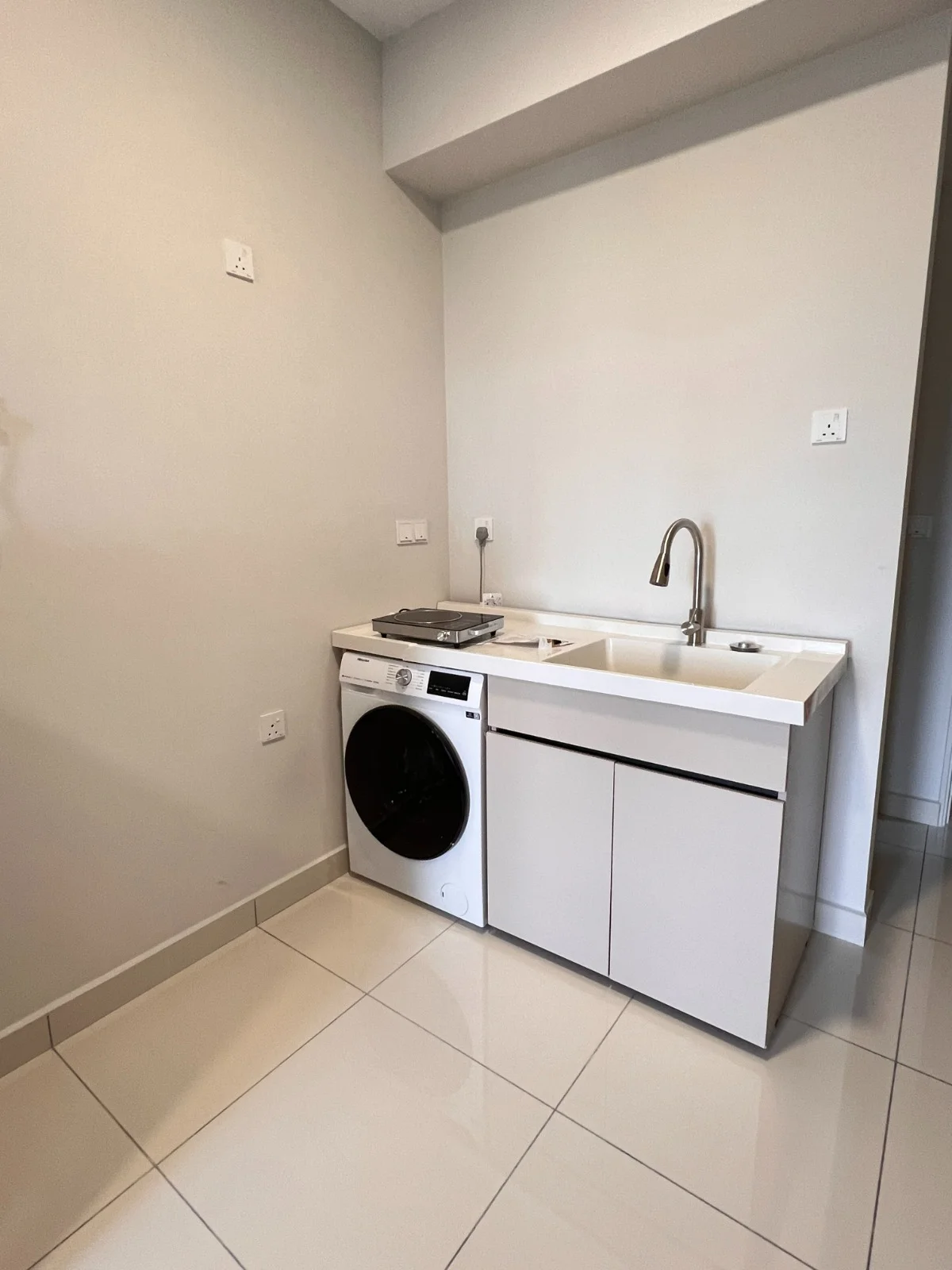 Laundry features: white cabinetry, single-bowl stainless-steel sink, chrome pull-down faucet, front-load washing machine in white, white countertop, white tiled flooring, light grey painted walls, wall-mounted electrical outlet; style: modern.