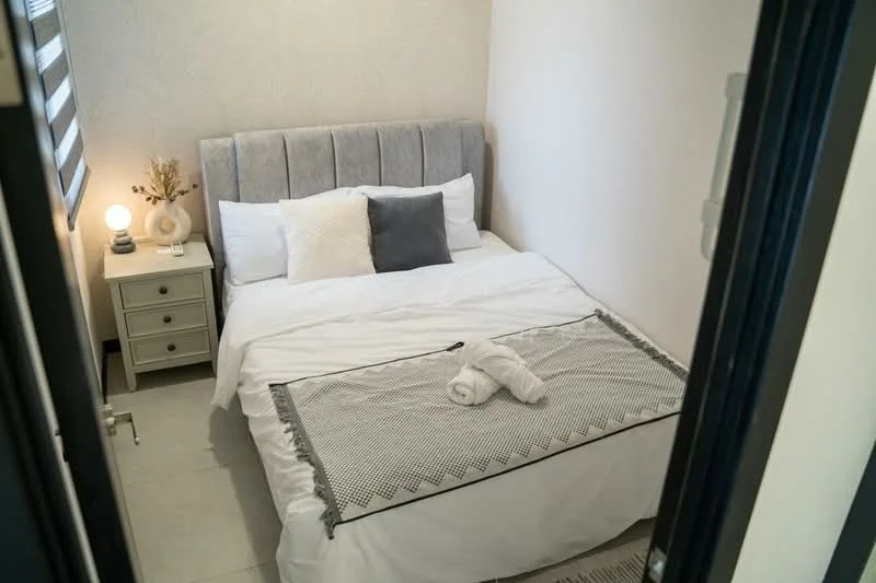 Bedroom features: queen-size bed with upholstered headboard, white bedding, decorative throw blanket, nightstand with drawer, table lamp, wall-mounted artwork, light grey accent wall, ceramic vase with dried flowers, sliding door, tile flooring; style: modern.