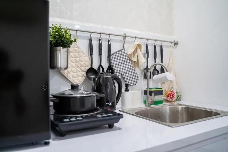 Kitchen features: white countertop, stainless-steel sink, chrome pull-down faucet, black electric kettle, black stovetop with 2 burners, black cooking pot, hanging pot holders, kitchen utensils on rack, green potted plant, checkered pot holder, cream pot holder, colorful dish sponge; style: modern.