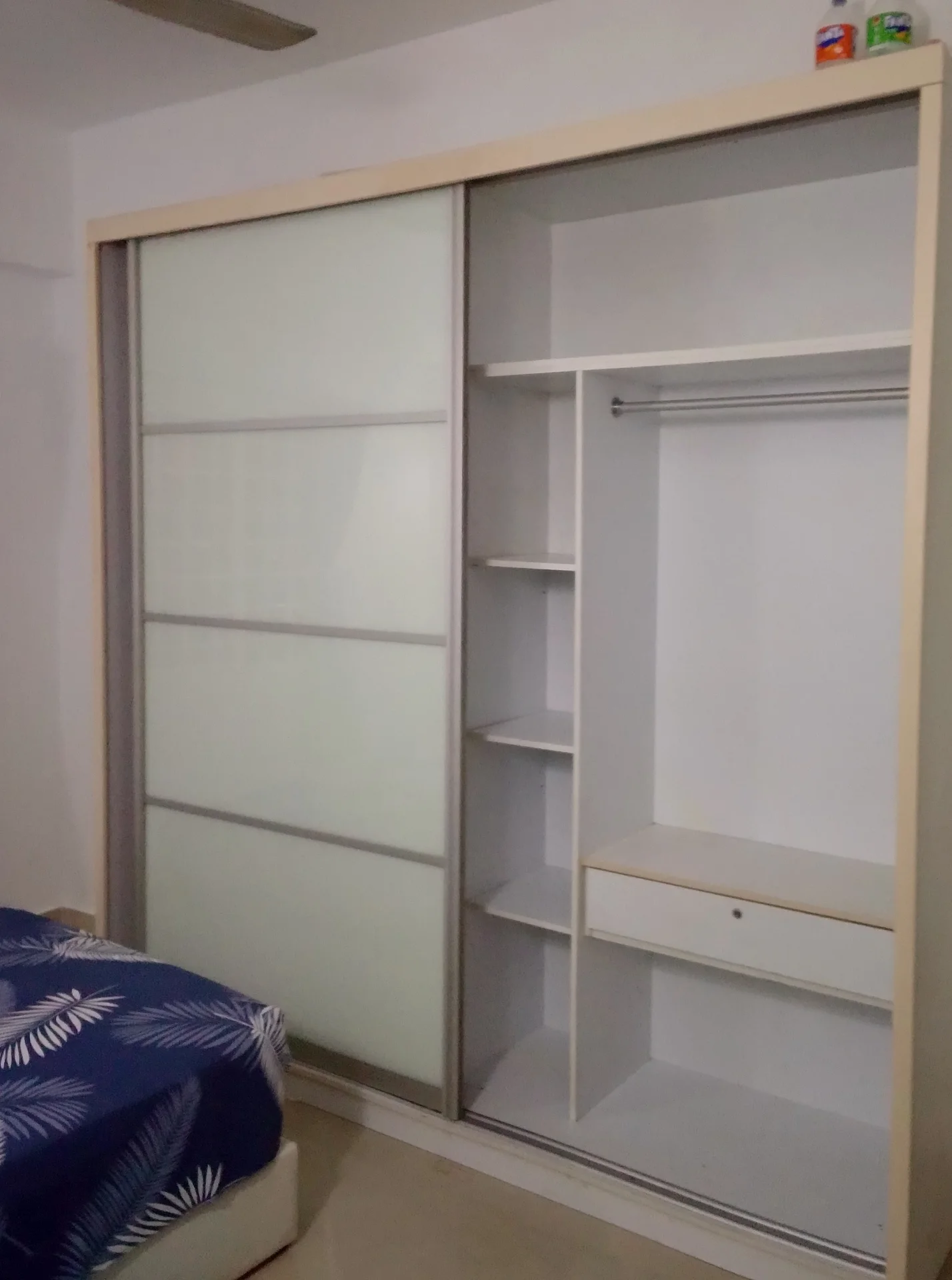 Room features: sliding wardrobe with frosted glass panels, built-in shelving, hanging rod, drawer unit, bed with patterned bedding, neutral wall colour, light-coloured flooring, ceiling fan; style: modern.