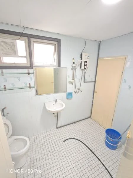 Bathroom features: white tiled walls, light blue painted walls, white ceramic sink, chrome basin mixer faucet, framed mirror, one-piece toilet, shower with handheld showerhead, white tiled flooring, glass shelf, blue plastic bucket, window for ventilation.