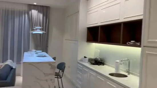 Kitchen features: white cabinetry with flat-panel doors, marble countertop, single-bowl stainless-steel sink, chrome pull-down faucet, built-in stove, under-cabinet lighting, open shelving, bar seating area, grey upholstered bar stool, light grey walls, curtains with a light fabric, recessed downlights; style: modern.