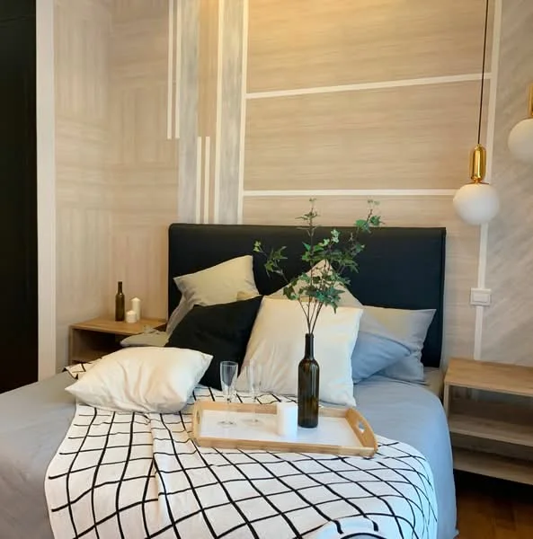 Bedroom features: king-size bed with upholstered headboard, decorative throw pillows in various colors, bedside tables with lamps, wooden tray on bed with decorative items, glass vase with greenery, white and black grid-patterned throw blanket, light wood flooring, wall with wood paneling, wall-mounted light fixture, neutral-colored bedding; style: modern.