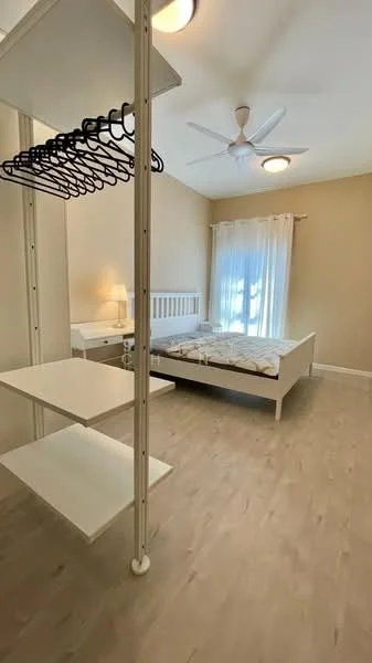 Bedroom features: queen-size bed, white bed frame, light wood flooring, ceiling fan, wall-mounted clothing rack, white shelving unit, sheer curtains, table lamp, window with natural light, neutral wall color; style: modern.