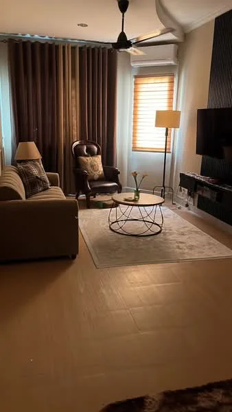 Living features: 3-seater fabric sofa in beige, accent chair in dark brown leather, round coffee table with metal base, area rug in light color, floor lamp with cream shade, wall-mounted TV, wooden TV console, curtains in dark brown, window with horizontal blinds, ceiling fan, decorative pillow on sofa, framed artwork on wall; style: modern.