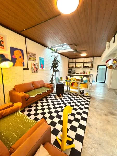 Living features: brown leather 3-seater sofa, brown leather 2-seater sofa, black and white checkered area rug, wooden ceiling with exposed beams, ceiling fan, large skylight, decorative wall art, green plant in pot, dining table with colorful chairs, open shelving unit, kitchenette area with countertop, wall-mounted storage, light fixture, decorative items on shelves; style: modern.