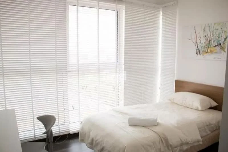 Bedroom features: single bed with white bedding, wooden headboard, floor-to-ceiling windows, white vertical blinds, light-colored walls, dark wood flooring, small white side table, decorative artwork on wall; style: modern; view: city.