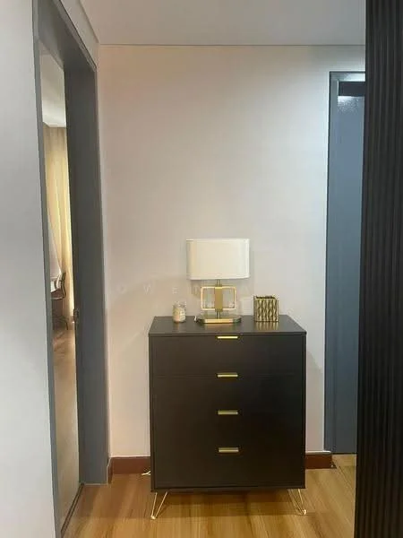 Room features: dark wood dresser with gold handles, table lamp with white shade, decorative gold geometric object, light-colored walls, wooden flooring, doorway leading to another room, doorway with dark frame, natural light from window; style: modern.