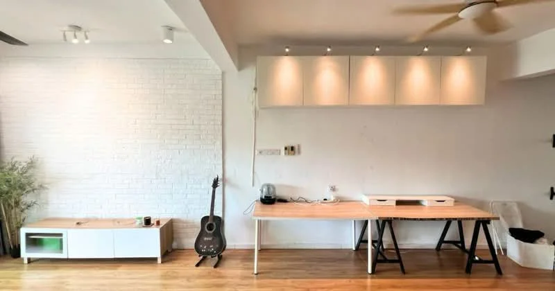 Room features: white painted walls, exposed brick accent wall, wooden flooring, ceiling fan, recessed downlights, light wood table, black metal legs on table, guitar, white console table, green accent light on console, cable management on wall, multiple wall-mounted cabinets; style: modern.