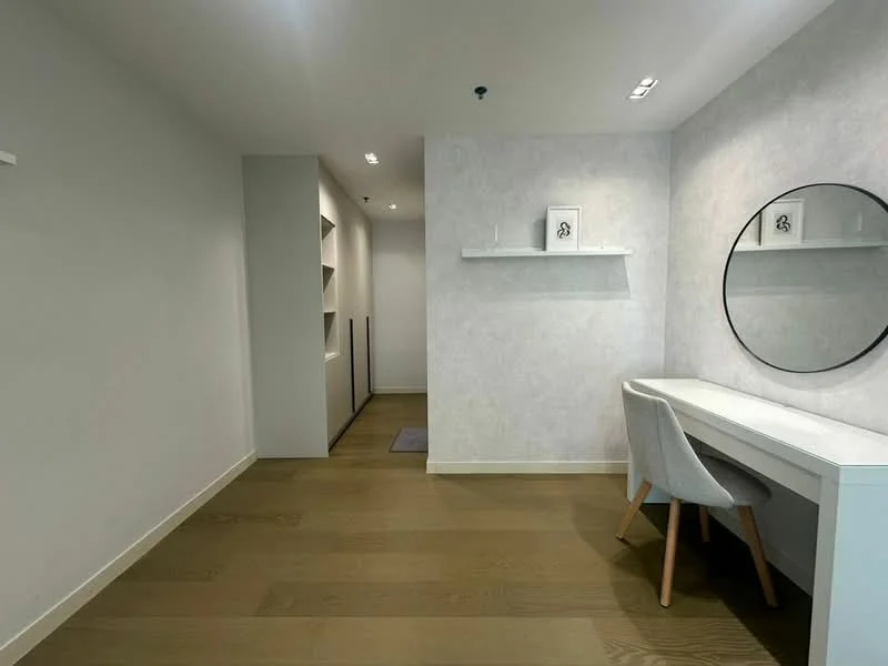 Room features: light wood flooring, white painted walls, wall-mounted shelf, round mirror, vanity table with drawers, white chair, recessed lighting, narrow hallway, built-in shelving unit; style: modern.