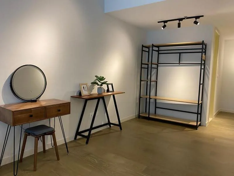 Room features: wooden flooring, white painted walls, track lighting, wooden console table with drawers, black metal shelving unit, round mirror, decorative plant, stool with wooden seat and metal legs; style: modern.