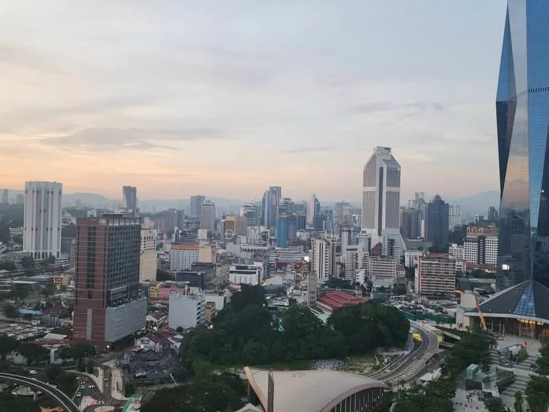 Exterior type: high-rise condominium; features: city skyline view, mixed-use urban environment, surrounded by greenery, variety of building heights, modern architectural designs, commercial and residential buildings, distant mountains in background, sunset lighting, urban landscape, roadways visible; style: modern.
