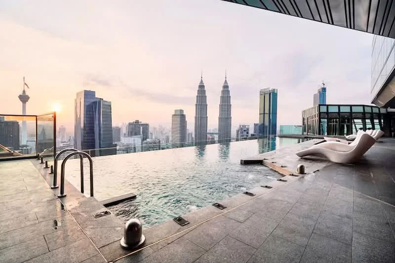 Exterior type: high-rise condominium; features: infinity pool, pool deck with loungers, glass railing, stone-paved flooring, city skyline view, surrounded by skyscrapers, sunset lighting; style: modern.