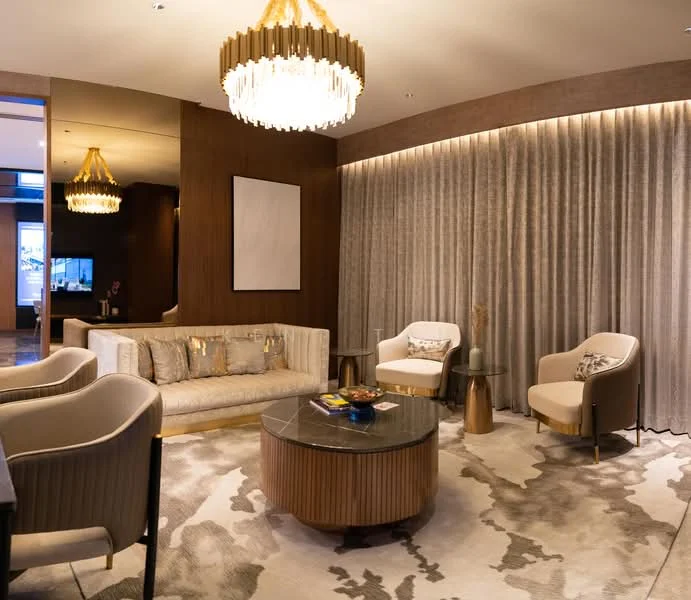 Living features: chandelier with crystal accents, sofa in light beige upholstery, accent pillows with metallic details, round coffee table with wooden base, area rug with abstract pattern, two accent chairs in white upholstery, curtains in light grey fabric, wall with wood paneling, recessed lighting, decorative wall art, side table with decorative items; style: modern.
