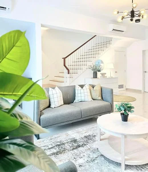 Living features: grey fabric sofa, decorative cushions with geometric patterns, round coffee table, area rug with abstract pattern, ceiling light fixture with multiple bulbs, wall-mounted air conditioning unit, staircase with wooden handrail, white walls, large window for natural light, indoor plants; style: modern.
