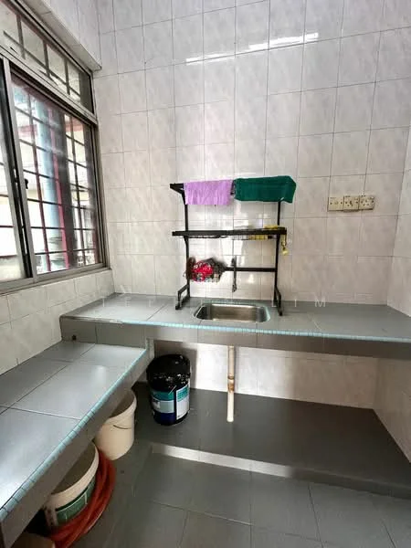 Kitchen features: grey laminate countertop, stainless-steel sink, wall-mounted shelf, white tiled walls, grey tiled flooring, window with metal grille, plastic buckets, cleaning supplies, dish rack, plumbing visible under sink.