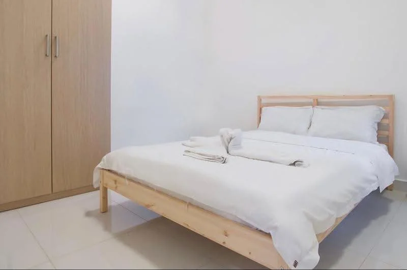 Bedroom features: queen-size bed with white bedding, wooden bed frame, light wood wardrobe, white painted walls, glossy tile flooring, two white pillows, folded towels on bed; style: minimalist.