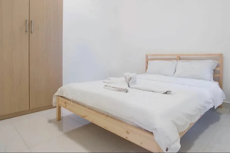 Bedroom features: queen-size bed with white bedding, wooden bed frame, light wood wardrobe with sliding doors, white painted walls, glossy tile flooring, two white towels on the bed; style: minimalist.