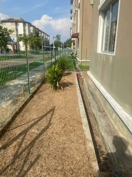 Exterior type: low-rise apartment block; features: landscaped garden with plants, fenced boundary, concrete pathway, grass area, sidewalk, two-storey building, windows with white frames, exterior wall in beige, red awning, paved area next to building; style: modern.