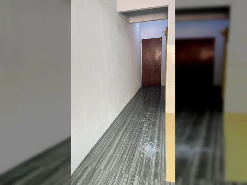 Hallway features: white painted walls, grey striped vinyl plank flooring, flat white ceiling, narrow corridor layout, brown wooden door, door frame opening, beige tiled wall section, recessed ceiling beam, plain wall surfaces; style: minimalist.