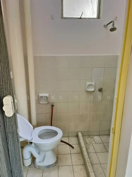 Bathroom features: white ceramic toilet, shower area with wall-mounted shower head, tile flooring, white tiled walls, drain in shower area, small window for ventilation, toilet paper holder, exposed plumbing, shower drain.