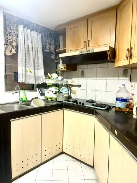 Kitchen features: L-shaped kitchen layout, wood cabinetry in light brown, black countertop, stainless-steel sink, chrome pull-down faucet, 4-burner gas hob, exhaust hood, white tiled backsplash, window with sheer curtains, dish rack, stacked plates and bowls, kitchen utensils on display.