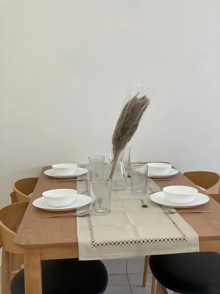 Dining features: wooden dining table, four wooden dining chairs, white ceramic plates, clear drinking glasses, table runner in beige, decorative dried flowers in vase, neutral wall color, tile flooring; style: modern.