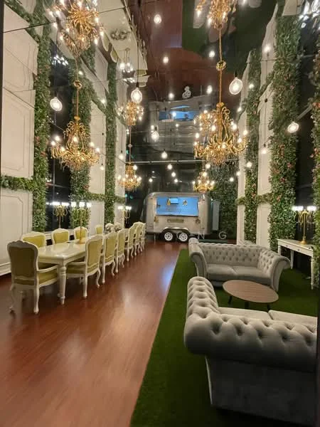 Living features: chandelier lighting, greenery wall decor, wooden flooring, sofa with tufted upholstery in grey, round coffee table, dining table with white and yellow upholstered chairs, artificial grass flooring, wall-mounted TV, decorative wall panels, exposed ceiling with hanging lights; style: modern.