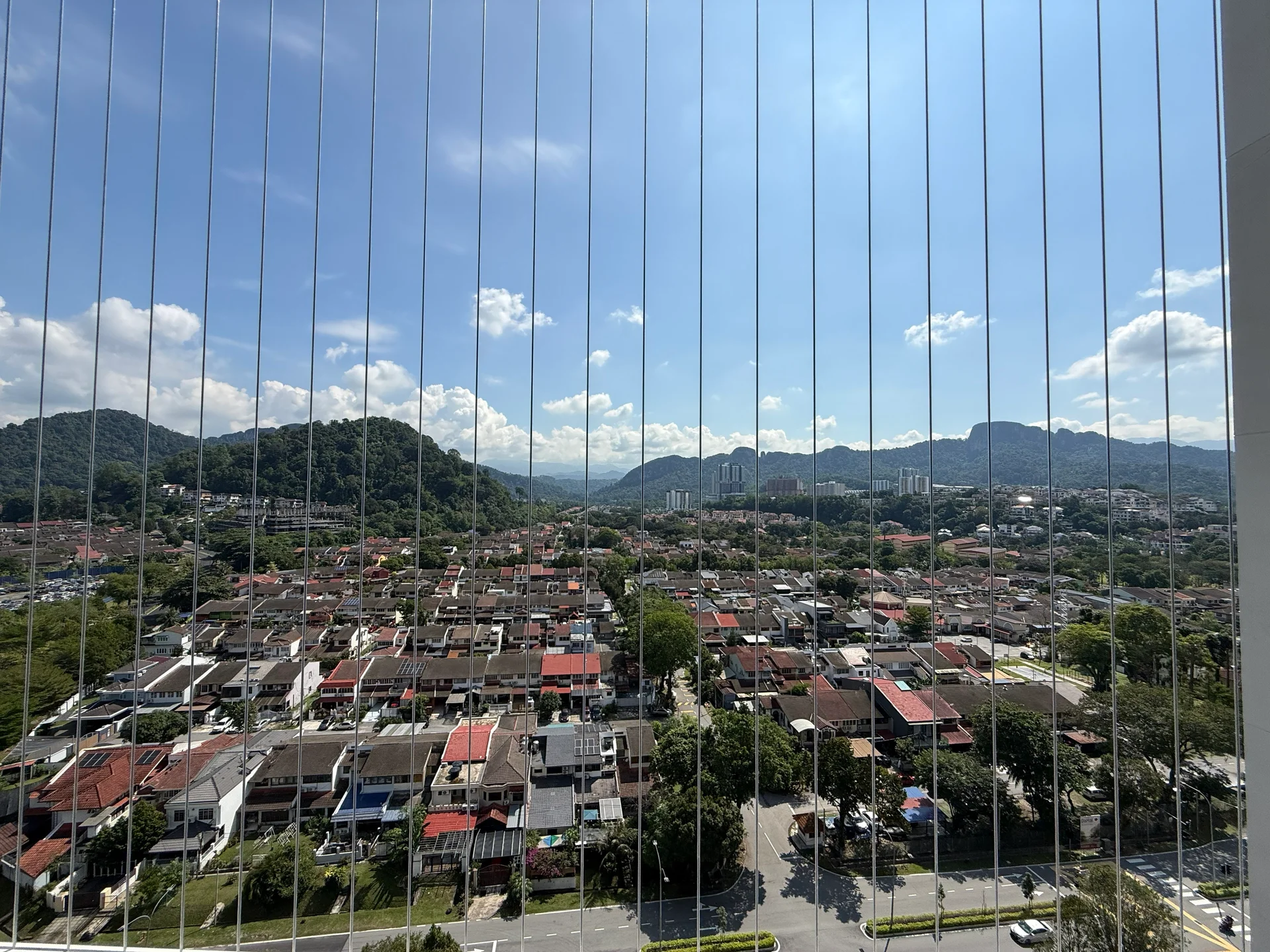 Exterior type: high-rise condominium; features: city skyline view, mountain view, surrounded by residential area, clear blue sky, greenery in the foreground, visible rooftops of houses, urban landscape, window with vertical blinds; style: modern.