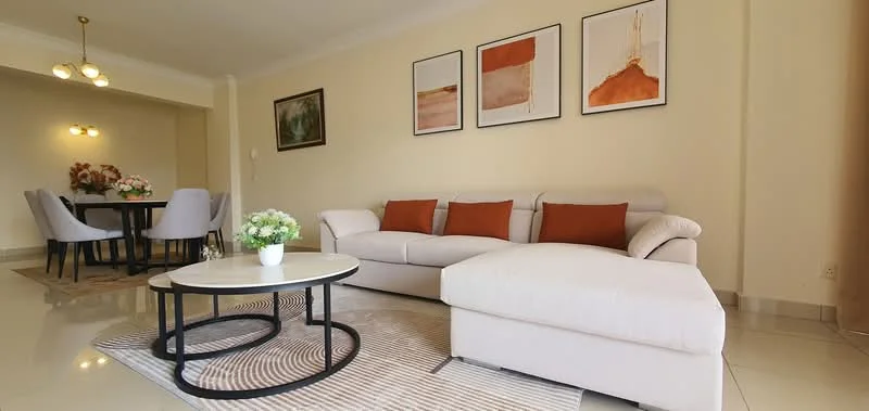 Living features: L-shape sectional sofa in light grey, orange accent cushions, round coffee table with black metal base, area rug with geometric pattern, framed artwork on walls, recessed ceiling lights, cream painted walls, tile flooring; style: modern. Dining features: round dining table, 4 upholstered dining chairs, floral centerpiece on table, cream painted walls, recessed ceiling lights; style: modern.