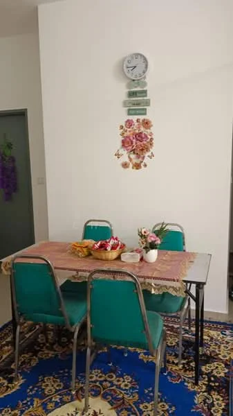 Dining features: dining table with tablecloth, four metal chairs with green upholstery, decorative wall clock, floral wall decor, blue area rug with floral pattern, flower vase on table, fruit basket on table, white walls.