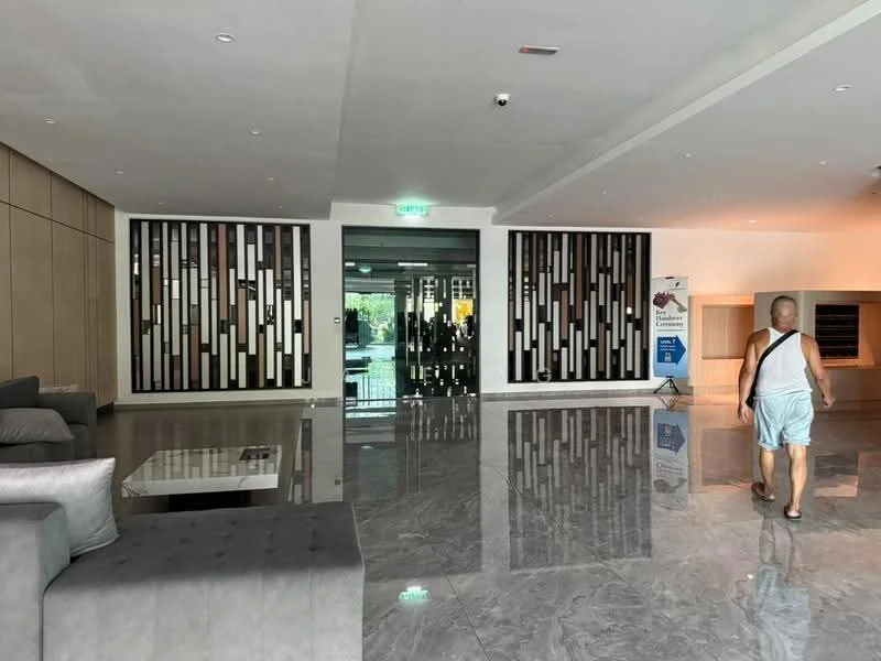 Room features: polished marble flooring, wood paneling on walls, glass entrance doors, recessed lighting, modern seating area with grey sofas, decorative wall with geometric patterns, large open space, elevators visible in background; style: modern.