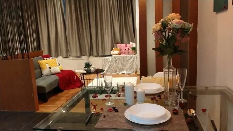 Dining features: glass dining table, white plates, wine glasses, decorative table runner, fresh flowers in vase, rose petals scattered on table, candles, wooden flooring; style: modern; view: living. Living features: grey fabric sofa, yellow and red throw pillows, decorative throw blanket, wall-mounted artwork, curtains, wooden accent wall, area rug; style: modern.