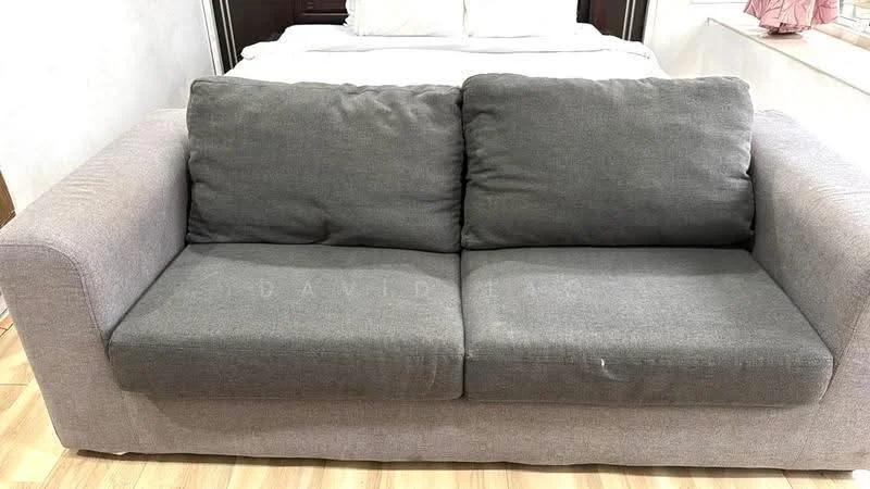 Room features: 2-seater fabric sofa in grey, wooden flooring, neutral wall colour, bed visible in background; style: modern.