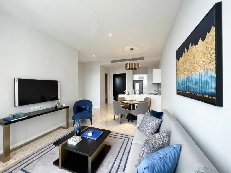 Living features: sofa in light grey fabric, blue accent chairs, coffee table with black top and gold legs, wall-mounted TV, decorative wall art with gold and blue colors, recessed downlights, area rug with geometric pattern, marble flooring, open layout to kitchen and dining area, dining table with four chairs visible in background; style: modern.
