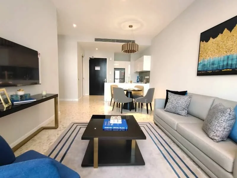 Living features: light grey leather sofa, blue throw pillows, glass coffee table, area rug with geometric pattern, wall-mounted TV, black console table, chandelier with gold accents, marble flooring, decorative wall art, dining area visible in background, open layout to kitchen, large windows with natural light; style: modern.