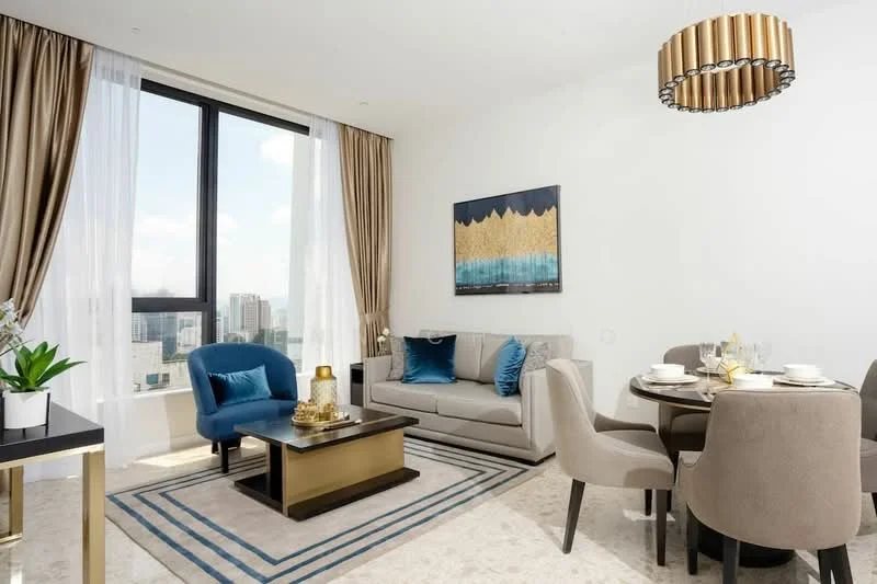 Living features: floor-to-ceiling windows, beige fabric sofa, decorative cushions in blue, coffee table with gold accents, area rug with geometric pattern, chandelier with circular design, wall-mounted artwork with gold and blue tones, dining table for four with upholstered chairs, sheer curtains, decorative plant in ceramic pot; style: modern; view: city.