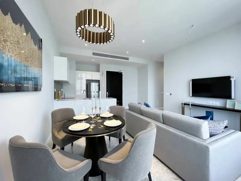 Dining features: round dining table, 4 upholstered dining chairs, chandelier with gold accents, decorative wall art with abstract design, white cabinetry, stainless-steel fridge, open kitchen layout, grey walls, recessed lighting, floor-to-ceiling windows, light-colored flooring; style: modern. Living features: 3-seater fabric sofa in light grey, wall-mounted TV, coffee table, decorative throw pillows, light grey walls, recessed lighting; style: modern. Kitchen features: white cabinetry, stainless-steel appliances, open layout, grey countertops, tile backsplash; style: modern.
