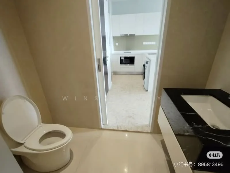 Bathroom features: one-piece toilet, single-sink vanity with black countertop, white sink, cream painted walls, marble tile flooring, recessed downlights, mirror above vanity, door leading to kitchen area; style: modern. Kitchen features: white cabinetry, built-in oven, freestanding range oven, microwave, countertop with backsplash, light-colored flooring, open layout to bathroom, recessed lighting; style: modern.
